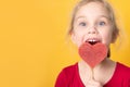 Cheerful perky little girl in a red dress holding a heart shaped lollipop.