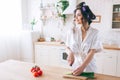 Cheerful positive young woman stand in kitchen and look at window. Cut green onion on desk. Female housekeeper wear