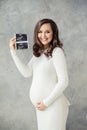 Cheerful pregnant woman showing ultrasound scan image and smiling on gray