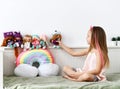 Cheerful pretty small girl with decorative bow and pink strands in hair sitting on bed and playing with collection of dolls