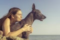 Cheerful pretty young girl sitting and hugging her dog xoloitzcuintli at the blue sky and sea on a stone beach Royalty Free Stock Photo