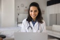 Cheerful pretty young Latin doctor woman enjoying work at laptop