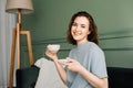 A cheerful pretty young woman with a cup of hot drink sitting on a couch enjoying a vacation. Couch Comfort. Cheerful Royalty Free Stock Photo