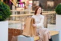 Cheerful pretty young woman sitting on bench with paper shopping bags at mall with modern interior, blurred background. Royalty Free Stock Photo