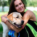 Cheerful pretty young woman sitting and hugging her dog at river bank in park Royalty Free Stock Photo