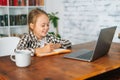 Cheerful primary child school girl doing homework writing notes in paper workbook sitting at table with laptop. Royalty Free Stock Photo