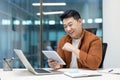 A cheerful programmer enjoys testing an application on a tablet, seated at a modern office desk with a laptop. Royalty Free Stock Photo