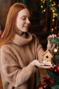 Cheerful red-haired young woman decorating Christmas tree beautiful toy at cozy living room. Royalty Free Stock Photo