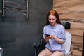 Cheerful redhaired young woman enjoying using phone sitting on toilet at bathroom, smiling looking to smartphone screen