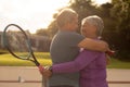 Cheerful romantic biracial senior couple embracing while standing at tennis court during sunset Royalty Free Stock Photo