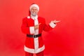 Cheerful santa claus in red costume pointing finger away showing freespace for advertising, looking at camera with toothy smile