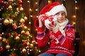 Cheerful santa helper girl with gift box sitting indoor near decorated xmas tree with lights, dressed in red sweater - Merry Royalty Free Stock Photo