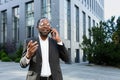 Cheerful and satisfied african american boss outside office building smiling and talking to colleagues on phone, man in Royalty Free Stock Photo