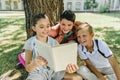 Cheerful schoolkids reading book while sitting under tree in park Royalty Free Stock Photo