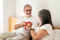 Cheerful senior caucasian woman gives gift box to husband, sit on bad in bedroom interior