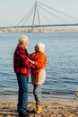 Cheerful senior citizens woman and man are standing and hugging on the lake, against the background of the bridge Royalty Free Stock Photo