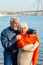 Cheerful senior citizens woman and man are standing and hugging on the lake, against the background of the bridge Royalty Free Stock Photo