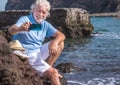 Cheerful senior man bare legs using smart phone for a selfie sitting on the rocks near the water enjoying the vacation in the sea Royalty Free Stock Photo