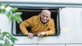 Cheerful senior man peering out from camper window holding glasses in hand. Joyful retirement road trip vibes Royalty Free Stock Photo