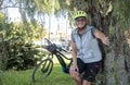 Cheerful senior sporty woman with electric bicycle and yellow helmet resting in shadow of tree in public park - active lifestyle