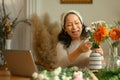 Cheerful senior woman having video call on laptop and arranging flowers in vase in living room. Retried lifestyle Royalty Free Stock Photo