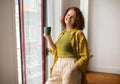 Cheerful senior woman holding green mug standing by window at home Royalty Free Stock Photo
