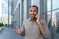 Cheerful and smiling African American office worker talking on the phone, man outside office building in shirt walking Royalty Free Stock Photo