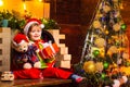 Cheerful smiling baby Santa boy holding his teddy bear and Christmas present near fire place and decorated New Year tree