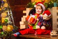 Cheerful smiling baby Santa boy holding his teddy bear and Christmas present near fire place and decorated New Year tree