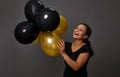 Cheerful smiling beautiful woman holding gold black shiny inflated air balloons against gray wall background with copy space. Royalty Free Stock Photo