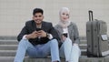 Cheerful smiling diverse business man and woman in hijab sitting on the stairs outdoors with smarphones and coffee