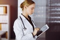 Cheerful smiling female doctor using tablet computer in sunny clinic. Portrait of friendly physician woman at work