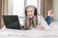 Cheerful smiling retired woman in headphones lying on bed at home, using laptop and making notes in her notepad, working Royalty Free Stock Photo