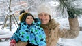 Cheerful smiling woman with baby son standing under tree branch and enjoying falling snow at winter Royalty Free Stock Photo