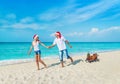 Cheerful smiling young couple in red Santa hats walking at tropical ocean sandy beach with sleds decorated fir-tree and golden fan