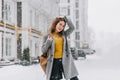 Cheerful smiling young woman in coat, with backpack walking in snowfall on street. Expressing true positive emotions