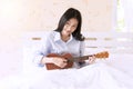 Young woman playing Ukulele relax on the white bed in the morning. Joyful girl play music with ukulele in bedroom Royalty Free Stock Photo