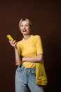 cheerful stylish blonde girl with bananas in string bag using yellow smartphone and smiling at camera