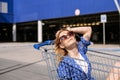 Cheerful stylish young woman in a blue dress on a sunny day sits in a shopping trolley in front of a supermarket Royalty Free Stock Photo