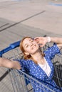 Cheerful stylish young woman in a blue dress on a sunny day sits in a shopping trolley in front of a supermarket Royalty Free Stock Photo