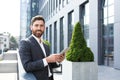 Cheerful and successful businessman works outdoors near the office holds a tablet, smiles reads good news Royalty Free Stock Photo