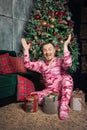 Cheerful surprised funny man in pink sleepwear with hands up sitting near decorated fir tree and Christmas present Royalty Free Stock Photo