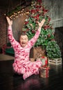 Cheerful surprised funny man in sleepwear with hands up sitting on the floor near decorated fir tree and Christmas present Royalty Free Stock Photo