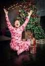 Cheerful surprised funny man in pink sleepwear with hands up sitting near decorated fir tree and Christmas present Royalty Free Stock Photo