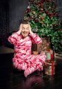 Cheerful surprised funny man in pink sleepwear sitting near decorated fir tree and Christmas present Royalty Free Stock Photo