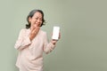 Cheerful and surprised 60s aged-asian woman palm touching chin, holding a smartphone