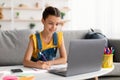 Cheerful teen sitting at desk, using laptop, drawing in copybook Royalty Free Stock Photo