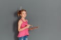 A cheerful teenage girl with a palette and brushes smiles against an empty gray wall. She will write a greeting and draw a picture