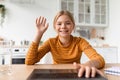 Cheerful teenage schoolgirl looks at laptop waving hand at table in kitchen interior