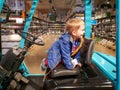 Portrait of cheerful toddler boy sitting on drivers seat of fork lifter in warehouse or big store with high shelves with